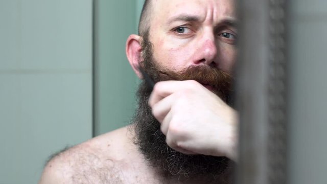 Bold man is combing his beard and mustache. Attractive man in bathroom looks in mirror and combes black hair on his face.
