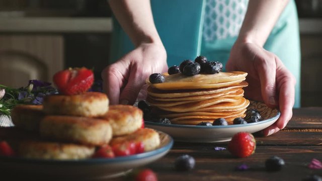 Female chief puts dish with fresh cocked blueberries and strawberries pancakes for breakfast in the morning light on a dark wooden table. Healthy Breakfast concept. Selective focus.