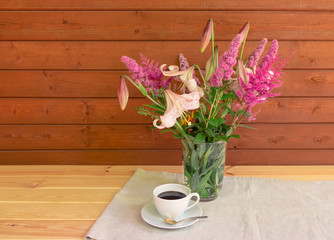 Bouquet of beige colored lilies with pink spots, pink spirea and astilbe flowers in glass  vase and cup of coffee on wooden table.