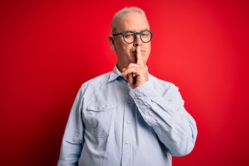 Middle age handsome hoary man wearing casual striped shirt and glasses over red background asking to be quiet with finger on lips. Silence and secret concept.