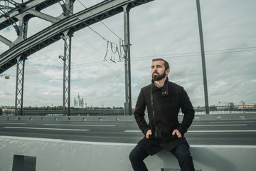 Dramatic look in a camera of a serious bearded handsome guy sitting on the curb stone of the bridge...