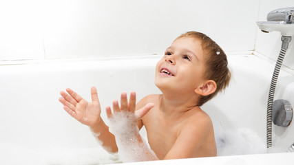 Portrait of cheerful smiling little boy enjoying washing in bath with soap foam