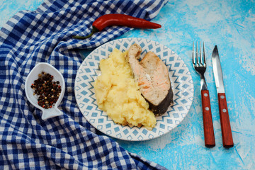 Mashed potatoes and red fish steak served on the plate on the blue background. Fork, knife,pepper and towel arranged near the plate. Homemade meal, tasty and nutritious. 