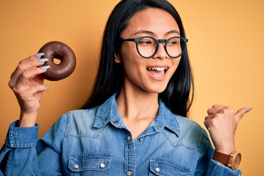 Young beautiful chinese woman holding chocolate donut over isolated yellow background pointing and showing with thumb up to the side with happy face smiling