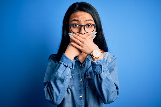 Young beautiful chinese woman wearing casual denim shirt over isolated blue background shocked covering mouth with hands for mistake. Secret concept.
