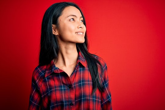 Young beautiful chinese woman wearing casual shirt over isolated red background looking away to side with smile on face, natural expression. Laughing confident.