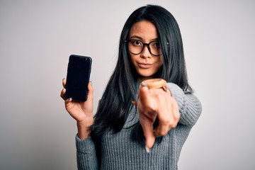 Young beautiful chinese woman holding smartphone over isolated white background pointing with...