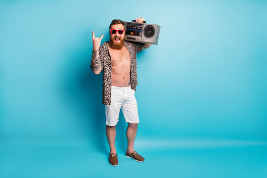 Full body photo of cool macho guy naked torso travel vacation hold retro tape recorder chill party show fingers horns wear shorts leopard shirt footwear sun specs isolated blue background