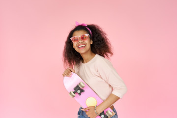Happy cool smiling young african american teen gen z girl skater wearing sunglasses holding skateboard looking at camera posing with skate in hands isolated on pink studio background, portrait.