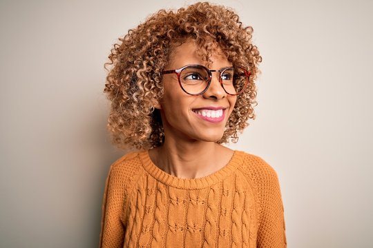 Young african american woman wearing casual sweater and glasses over white background looking away to side with smile on face, natural expression. Laughing confident.