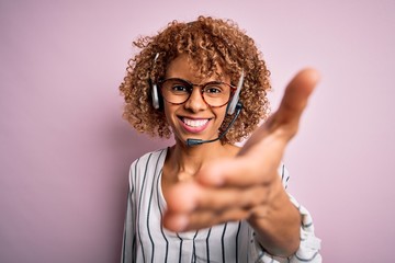 African american curly call center agent woman working using headset over pink background smiling friendly offering handshake as greeting and welcoming. Successful business.