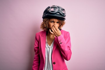 African american motorcyclist woman with curly hair wearing moto helmet over pink background smelling something stinky and disgusting, intolerable smell, holding breath with fingers on nose. Bad smell