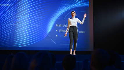 On-Stage Successful Female Speaker Presents Technological Product, Uses Remote Control for Presentation, Showing Infographics, Statistics Animation on Big Screen. Live Event / Business Conference