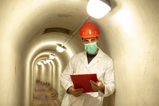 Tecnico con caschetto protettivo e camice bianco con n mano una coratella con dei documenti  dentro un tunnel sotterraneo