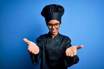 Young african american chef woman wearing cooker uniform and hat over blue background smiling cheerful offering hands giving assistance and acceptance.