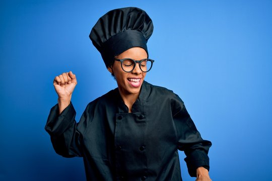 Young african american chef woman wearing cooker uniform and hat over blue background Dancing happy and cheerful, smiling moving casual and confident listening to music