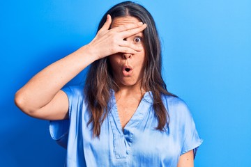 Young beautiful brunette woman wearing casual t-shirt standing over isolated blue background peeking in shock covering face and eyes with hand, looking through fingers afraid