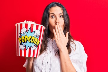 Young beautiful brunette woman eating pack of popcorns over isolated red background covering mouth...