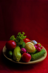 Vegetables on a wooden plate: potatoes, cucumbers, tomatoes, radishes, onions, garlic, herbs, zucchini, lettuce on a red background