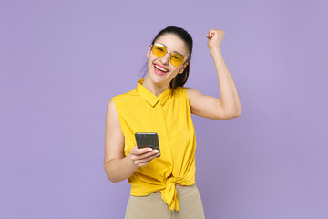 Happy young brunette woman girl in yellow casual shirt posing isolated on violet wall background studio portrait. People lifestyle concept. Mock up copy space. Using mobile phone doing winner gesture.