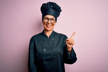 Middle age brunette chef woman wearing cooker uniform and hat over isolated pink background with a...