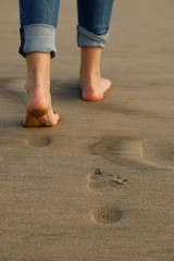 A woman wearing jeans walking on the beach sand and leaving footprints behind her. low section and close up view of the girl's feet. Vertical background concept.