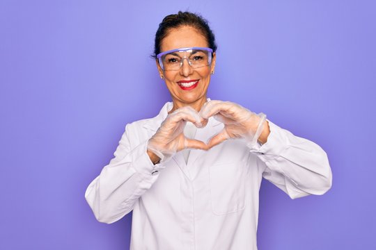 Middle age senior scientist woman wearing coat and laboratory glasses over purple background smiling in love showing heart symbol and shape with hands. Romantic concept.