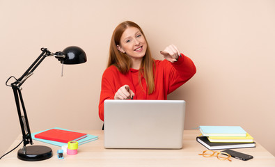 Young student woman in a workplace with a laptop points finger at you while smiling