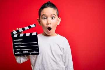 Young little boy kid filming video holding cinema director clapboard over isolated red background...