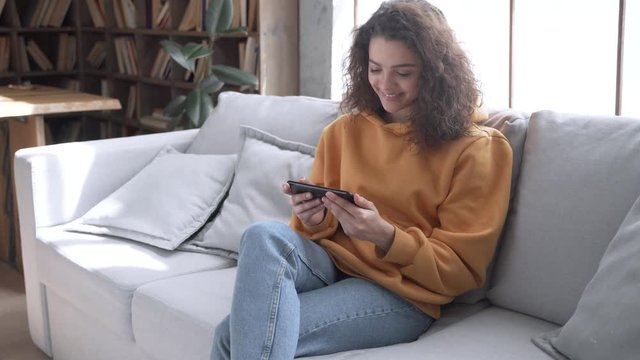Happy young hispanic latin teen girl sit on sofa at home holding phone looking at mobile screen talking video calling distance friend online in mobile chat app using smartphone videochat application.