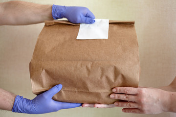 Male hands in blue latex glove gives paper bag to female hands. Closeup of male hands in gloves hold brown craft paper bag with blank white sticker. Delivery service concept. Copy space area.