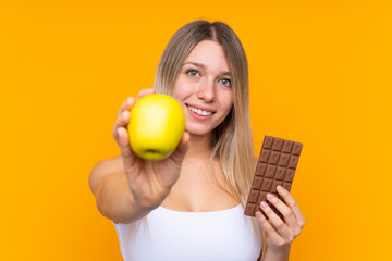 Young blonde woman over isolated blue background taking a chocolate tablet in one hand and an apple...