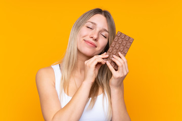 Young blonde woman over isolated blue background taking a chocolate tablet and happy