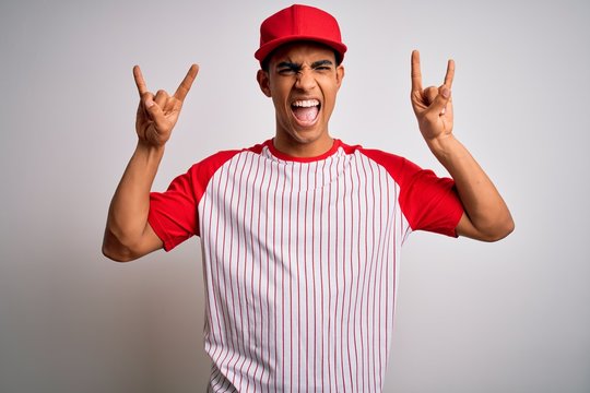 Young handsome african american sportsman wearing striped baseball t-shirt and cap shouting with crazy expression doing rock symbol with hands up. Music star. Heavy music concept.