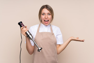 Young blonde woman using hand blender with shocked facial expression