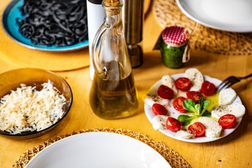 Plates with salad caprese, oil, grated cheese and black spaghetti on table.