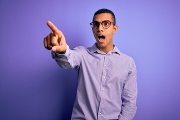 Handsome african american man wearing striped shirt and glasses over purple background Pointing with finger surprised ahead, open mouth amazed expression, something on the front