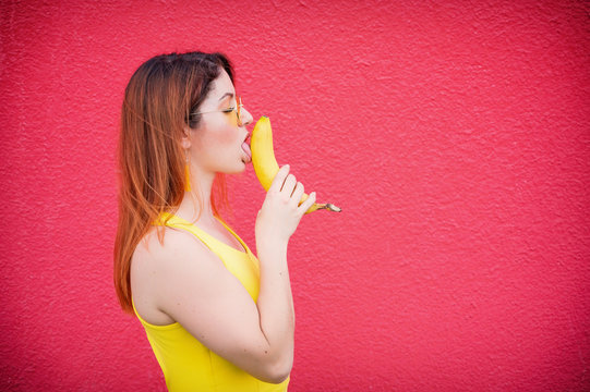 Beautiful red-haired woman in a yellow dress and glasses kisses a banana on a red background. Portrait of a girl fantasizing with fruit.