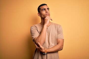 Young handsome african american man wearing casual t-shirt standing over yellow background with hand on chin thinking about question, pensive expression. Smiling with thoughtful face. Doubt concept.