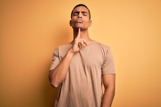 Young handsome african american man wearing casual t-shirt standing over yellow background Thinking concentrated about doubt with finger on chin and looking up wondering