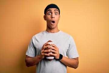 Young handsome african american man holding rugby ball over isolated yellow background scared in...