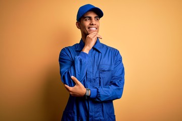 Young african american mechanic man wearing blue uniform and cap over yellow background with hand on chin thinking about question, pensive expression. Smiling with thoughtful face. Doubt concept.