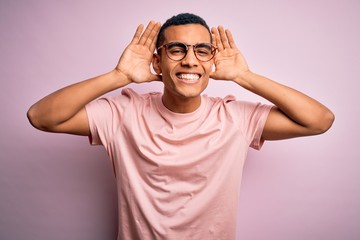 Fototapeta na wymiar Handsome african american man wearing casual t-shirt and glasses over pink background Trying to hear both hands on ear gesture, curious for gossip. Hearing problem, deaf