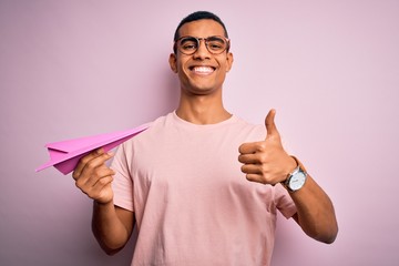 Young handsome african american man holding paper airplane over isolated pink background happy with...