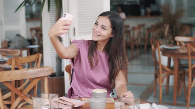 Brunette woman sits in outdoor cafe, drinks coffee and takes selfie on phone