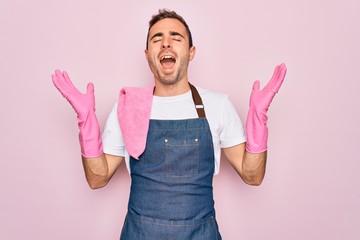 Young cleaner man with blue eyes cleaning wearing apron and gloves over pink background celebrating...