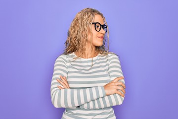 Fototapeta premium Beautiful blonde woman wearing casual striped t-shirt and glasses over purple background looking to the side with arms crossed convinced and confident