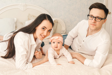 Happy young father, mother and cute baby girl lying on light bed.