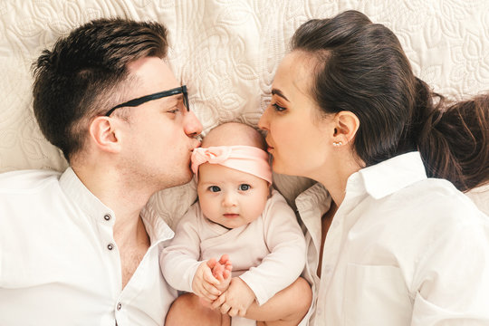 Happy young father, mother and cute baby boy lying on light bed. Mother and father kiss baby.