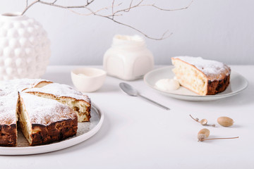 Homemade simple apple cake or pie sliced and sprinkled powdered sugar on grey concrete table decorated dry twig in vase. Autumn or winter treat. Fika. Hygge